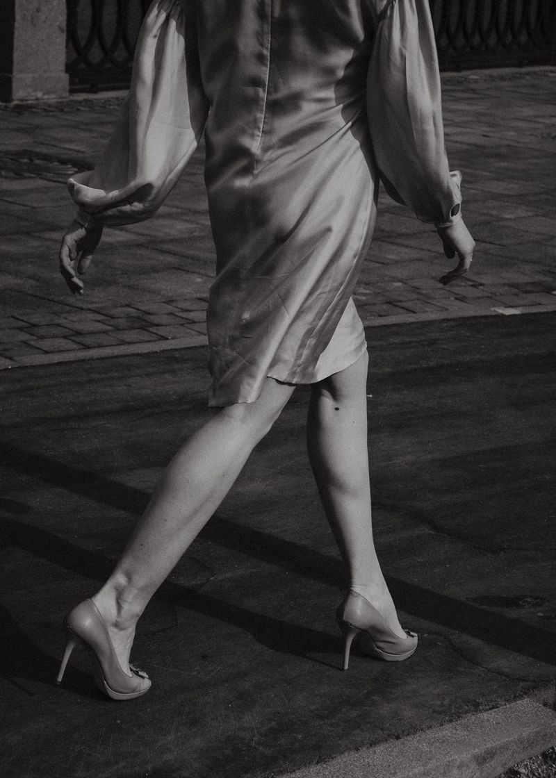 A woman in a silk dress walking across a sunlit city street.